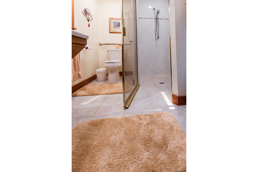 A shower door swings open to reveal a threshold-free shower that someone in a wheelchair could enter without difficulty.  Several peach bathmats decorate the floor, and matching towels hang on a bar in the bathroom. Installed grab bars around the toilet are visible.
